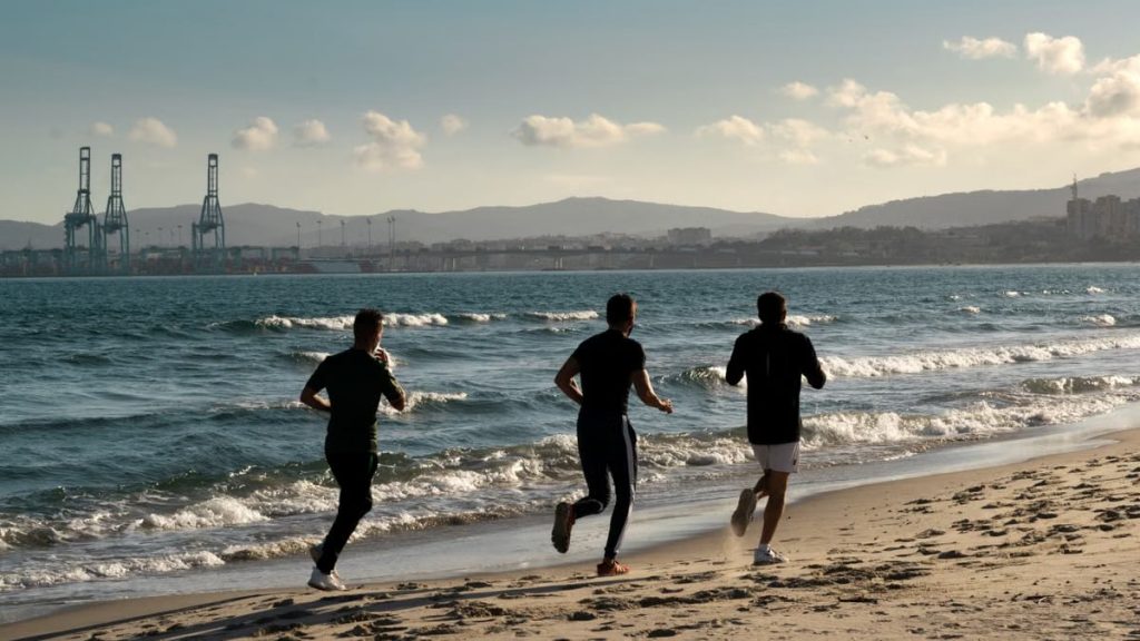 Three men jogging along a scenic beach, embracing a healthy lifestyle under a clear sky.