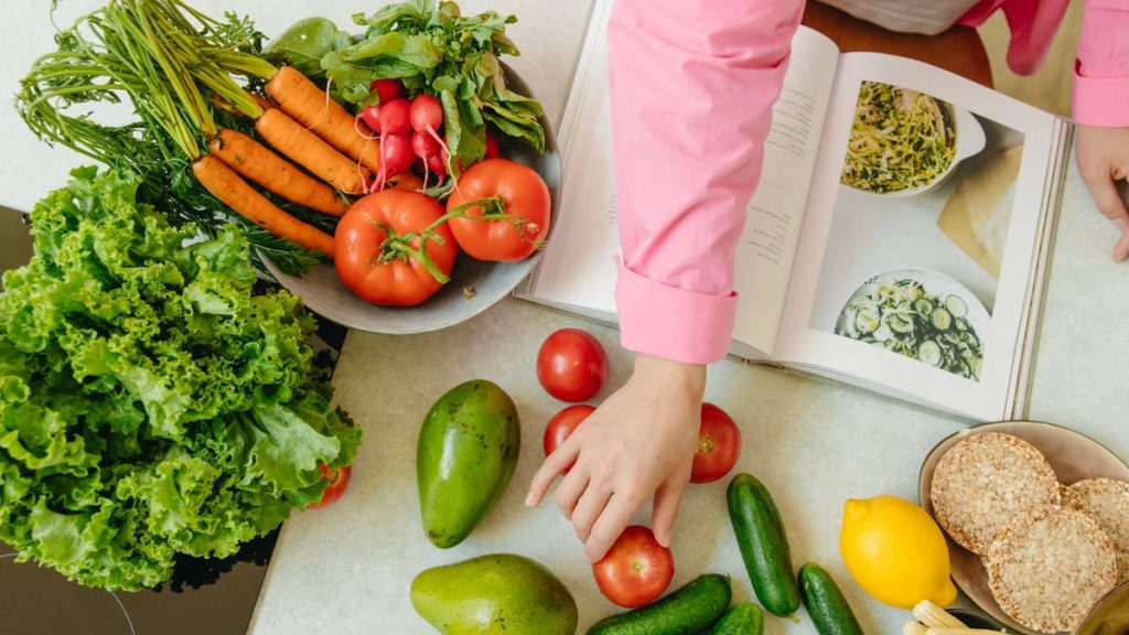 Top view of fresh vegetables and hands arranging them with a recipe book, promoting healthy eating.
